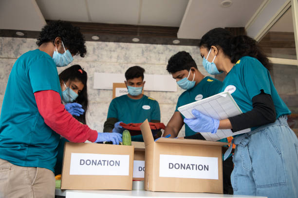 Low angle view, group of volunteers busy working by arranging vegetables and clothes on donation boxes and noting down during coronavirus covid-19 pandemic lockdown for needy people